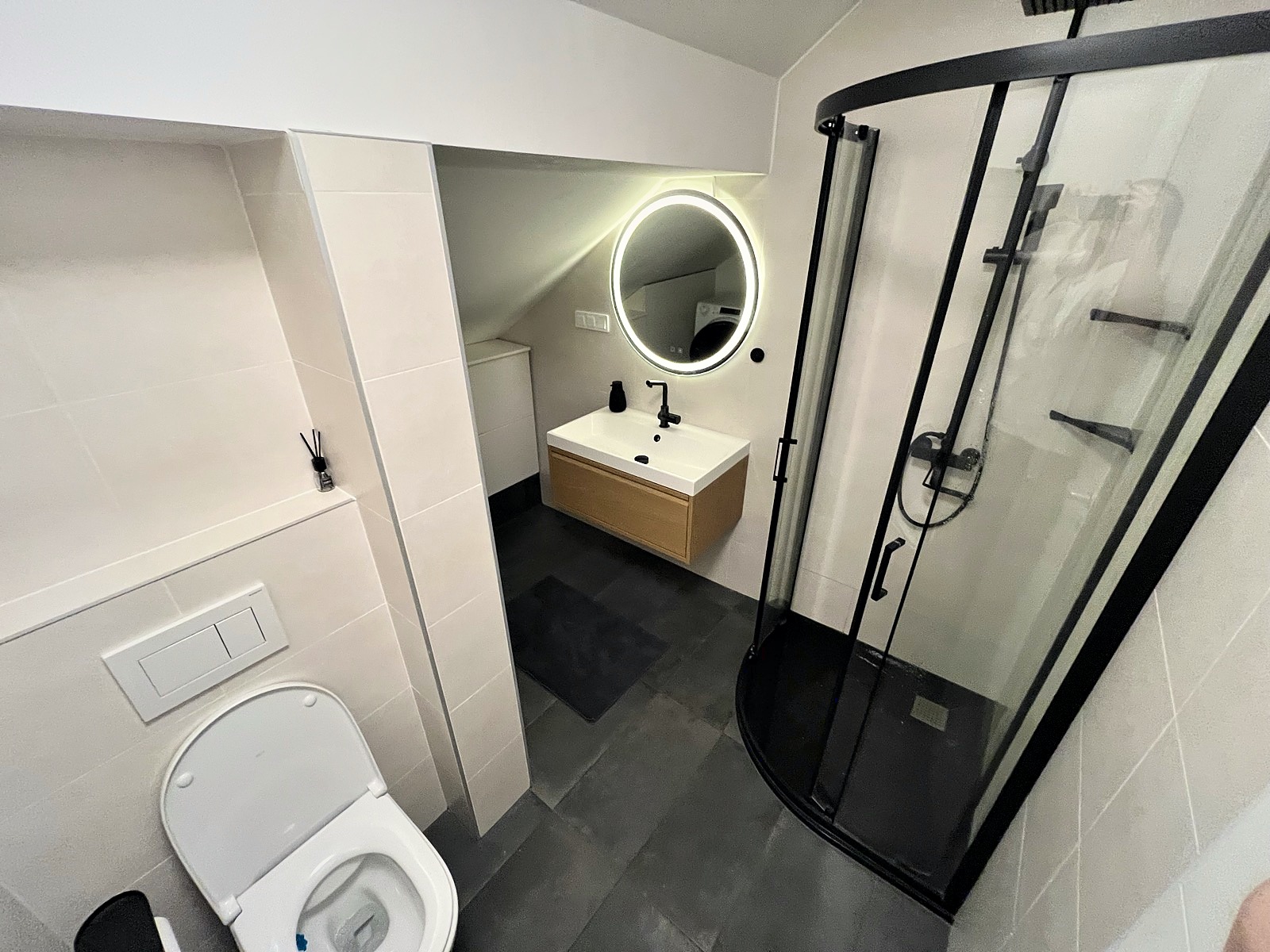 Modern bathroom with curved black-frame shower, illuminated circular mirror, and slate floor